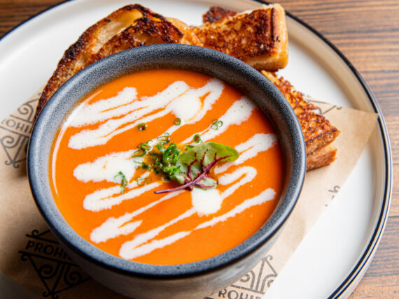 A bowl of tomato soup with a drizzle of white cream. Slices of grilled cheese are on a plate around the soup.