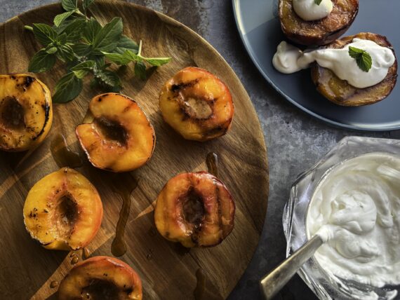 Grilled peaches on a wooden platter with a Bourbon and Brown-Sugar Butter Glaze, whipped cream, and baby basil leaves