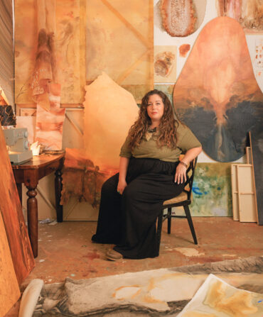 A woman sits in a chair in an art studio surrounded by earthy paintings