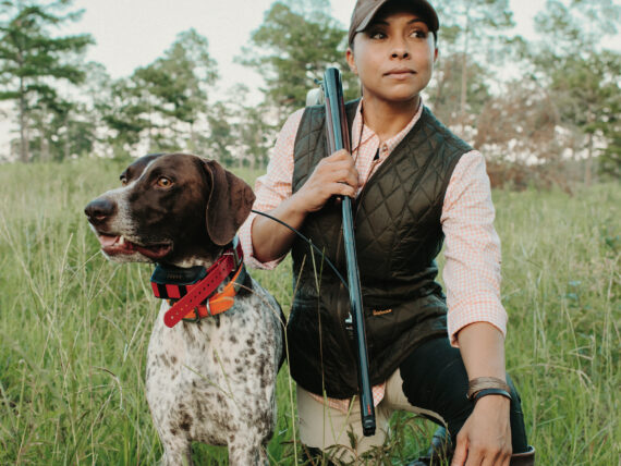 A woman kneels in a grassy field with her dog, a shorthaired pointer with white and brown fur, to the left