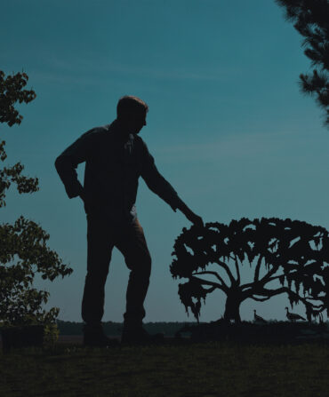 A man stands at dusk with a metal sculpture of a cut-out tree