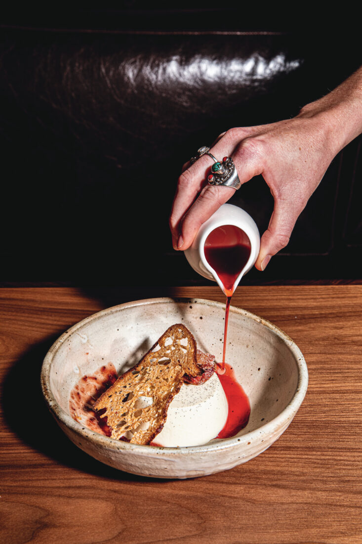 A hand pours red jammy liquid over panna cotta in a bowl