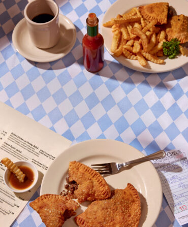 two plates of food on a blue and white checkered tablecloth with fries, ketchup, hot sauce and a cup of coffee